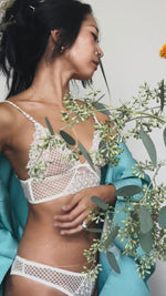 Woman wearing a white lace bra and panties with a light blue kimono and greenery in the background.